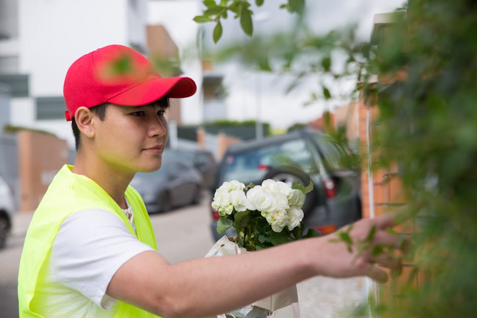 A young male florist in a red cap and high-visibility yellow vest is outdoors, arranging a bouquet of white flowers with lush green foliage. He is holding the bouquet in one hand and carefully adjusti