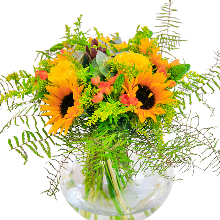 A first glance at this captivating bouquet conjures up the golden glow of summer evenings in Richmond-perhaps after a day spent exploring the floral wonders of Kew Gardens or meandering through Richmond Green. Leading the arrangement, generous sunflowers radiate outward with bright yellow petals wrapped around distinctive dark centers, filling the space with energy and optimism. Nestled between these bold blooms, classic yellow roses unfold layer upon layer of softly spiralled petals, their texture reminiscent of silk and their hue echoing the mellow light at golden hour. Piercing through the yellows, glossy clusters of red hypericum berries create vibrant contrast, their presence a playful nod to the colourful stalls found at Richmond's artisan markets. Wisps of green-fanned fern fronds and sprigs of delicate goldenrod, topped with minute yellow blossoms-bring a sense of airiness and gentle motion, while silvery eucalyptus leaves add a subtle touch of woodland calm. The clear, rounded glass vase gracefully displays the slender green stems, immersed in fresh water-a detail that enhances the sense of vitality and celebration intrinsic to the design. Meticulously arranged with attention to harmony and flow, this bouquet is well-suited for special celebrations, heartfelt gifts, or simply infusing a Richmond home with seasonal brightness and a sense of community connection.