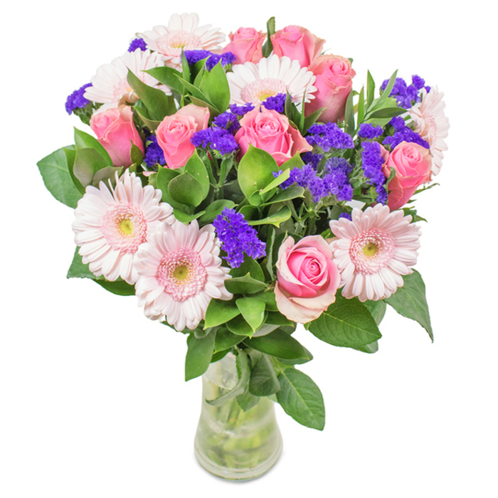 Positioned against a crisp white background, this sophisticated Richmond bouquet is presented in a sleek, clear glass vase that displays each element with clarity and intention. At its heart are approximately 7 to 9 medium and soft pink roses, their petals either tightly rolled or gently beginning to reveal a classic, spiral pattern symbolic of enduring affection. Interspersed are 5 to 7 elegant pale pink gerbera daisies, easily identified by their broad, round heads and myriad slender petals fading to nearly white at the centres. Vivid clusters of purple statice lace throughout the arrangement, providing a burst of color and rich texture that evokes the vibrant stalls of Richmond's weekly markets. The deep green, dewy foliage encircles the entire composition, giving it body and an inviting freshness reminiscent of shaded glades near Old Deer Park. The transparent vase reveals fresh-cut stems immersed in water, evoking the feeling of a locally sourced bouquet ready for delivery. Ideal for birthdays, anniversaries, Mother's Day, or simply adding a charming accent to a home or office overlooking Richmond Bridge, this floral ensemble radiates joyful celebration and heartfelt affection.